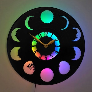 May include: A black vinyl record clock with a rainbow-colored clock face and eight moon phases around the edge. The moon phases are lit up in different colors.