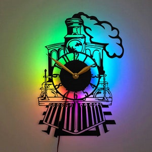 Puede incluir: Un reloj de pared de metal negro con forma de locomotora con una esfera y agujas doradas. El reloj está iluminado con luces de colores arcoíris.