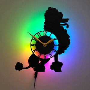 Op de afbeelding: Een zwart silhouet van een poedel met een regenboogklok in het midden. De wijzers van de klok zijn goud en de wijzerplaat heeft zwarte cijfers.
