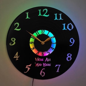 Peut inclure: Une horloge en vinyle noir avec un anneau lumineux LED arc-en-ciel autour du centre. Le cadran de l'horloge a des chiffres blancs et une aiguille des heures et des minutes dorée. Le texte "We're All Mad Here" est imprimé sous le cadran de l'horloge.