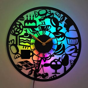 Può includere: Un orologio bianco e nero con una luce LED arcobaleno dietro. Il quadrante dell'orologio è decorato con immagini di dolci, torte e altre leccornie.