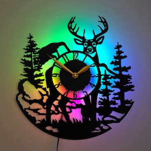 May include: A black silhouette of a deer with antlers standing in a forest scene. The deer is in front of a clock face with a rainbow colored background. The clock face has gold hands and black Roman numerals.
