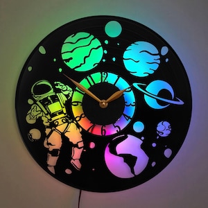 May include: A black vinyl record clock with a colorful astronaut design. The clock features a rainbow-colored clock face and a silhouette of an astronaut floating in space with planets and stars. The clock is illuminated with LED lights.
