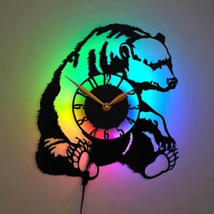 May include: A black bear silhouette clock with a rainbow-colored clock face. The clock hands are gold and the bear is holding the clock face in its paws.
