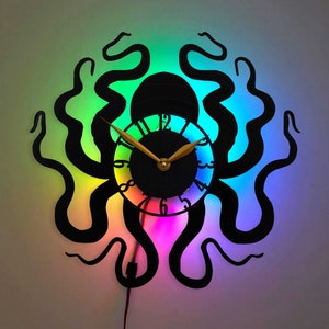 Op de afbeelding: Een zwarte octopus wandklok met een regenboog LED-licht. De wijzerplaat van de klok heeft een zwarte achtergrond met witte cijfers en gouden wijzers.