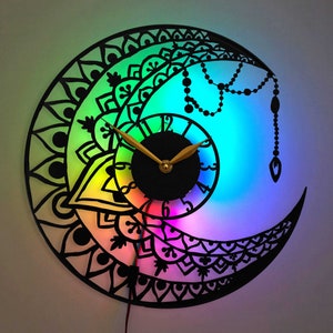 May include: A rainbow-colored clock with a black mandala design. The clock is shaped like a crescent moon and has a gold-colored hour and minute hand.