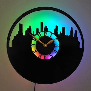 May include: A black vinyl record clock with a cityscape silhouette and a rainbow-colored clock face. The clock has gold hands and a black background.
