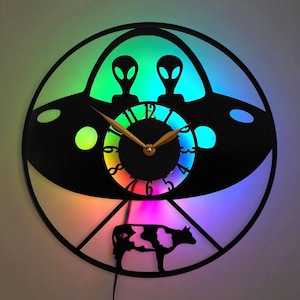 May include: A black vinyl record clock with a rainbow-colored LED light. The clock features a silhouette of a UFO with two aliens inside and a cow underneath. The clock face is a circular design with numbers 1-12 and a gold-colored hour and minute hand.