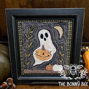 Könnte beinhalten: Ein gerahmter Halloween-Teppich mit schwarzem Rahmen. Der Teppich zeigt einen weißen Geist, der einen orangefarbenen Kürbis, einen Halbmond und zwei kleine Kürbisse hält. Der Hintergrund ist dunkelblau mit grünen und braunen Akzenten. Das Logo The Bonny Bee befindet sich unten rechts.
