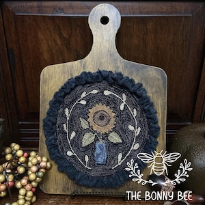May include: Wooden cutting board with a dark blue, circular, embroidered design featuring a sunflower in a blue vase, surrounded by a white leaf design. The design is framed by a dark blue, ruffled edge. The Bonny Bee logo is in the lower right corner.
