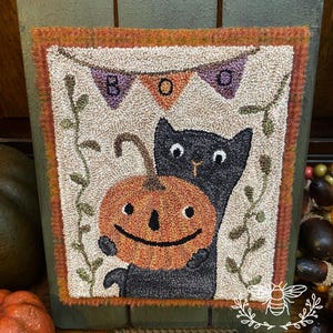 Könnte beinhalten: Ein handgefertigter Halloween-Teppich mit einer schwarzen Katze, die einen orangefarbenen Kürbis mit einem lächelnden Gesicht umarmt. Der Teppich hat ein Banner mit der Aufschrift "BOO" und ist in einem grünen Holzrahmen gefasst. Das Logo The Bonny Bee befindet sich unten.