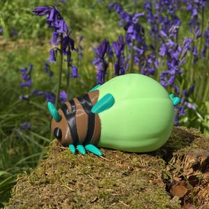 A direct, head-on view of the 3D printed Palia Lantern Bug collectible on a light wood surface. This symmetrical composition highlights the details of the bug's face, including the black eyes, brown head structure, and the four turquoise antennae. The black-and-brown striped body segments recede behind the head, with turquoise legs fanned out on both sides. The large, round, light-green abdomen is centered at the back, all against a blurred blue background.