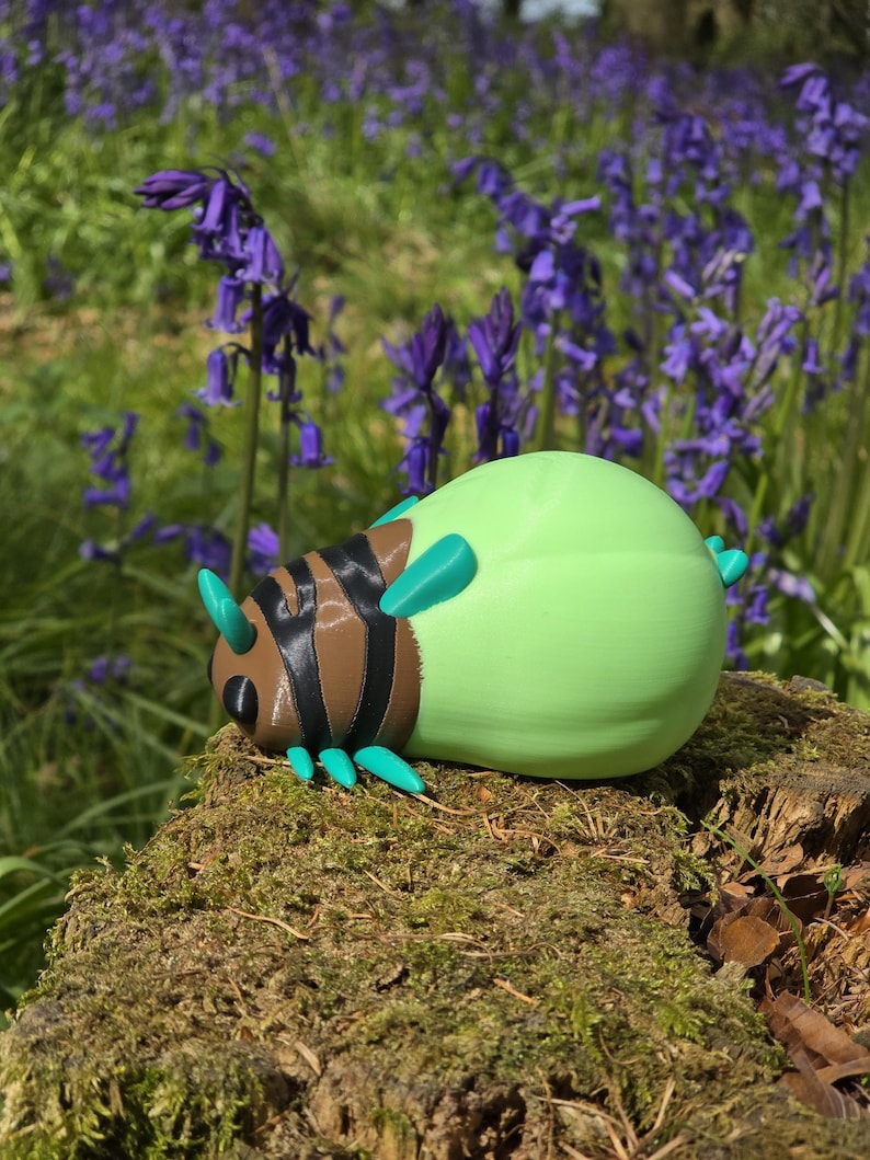 A direct, head-on view of the 3D printed Palia Lantern Bug collectible on a light wood surface. This symmetrical composition highlights the details of the bug's face, including the black eyes, brown head structure, and the four turquoise antennae. The black-and-brown striped body segments recede behind the head, with turquoise legs fanned out on both sides. The large, round, light-green abdomen is centered at the back, all against a blurred blue background.