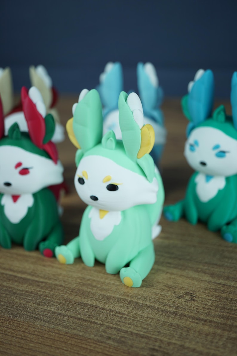 An angled close-up shot focused on the Spring Flutterfox miniature, featuring its mint green, white, and yellow colors. It sits on a wooden table, with other blurred Flutterfox figures, including the teal Summer fox, flanking it.