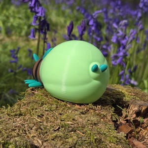 A detailed high-angle shot focused on the massive, light-green glowing abdomen of the 3D printed Palia Lantern Bug collectible. This view, taken from behind and slightly above, highlights the smooth, uniform color of the abdomen and the two small turquoise bumps on its surface. The head and striped body segments, with their turquoise legs and antennae, are visible and receding to the right, all set against a light wood surface and a blurred blue background.