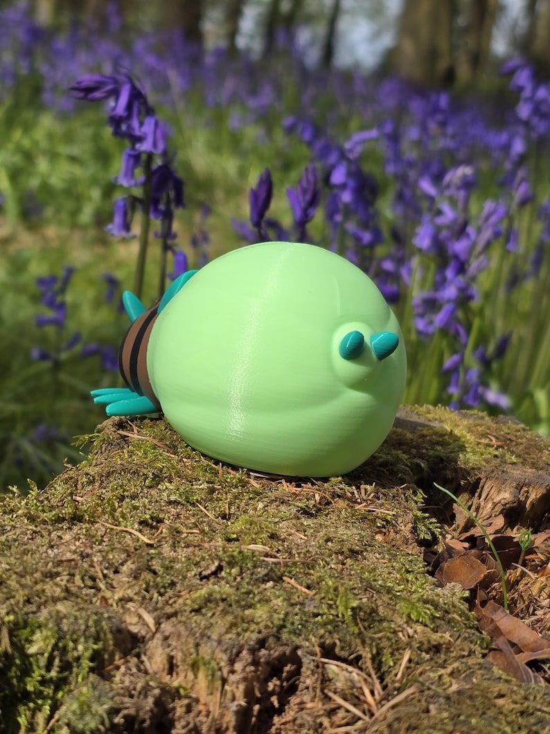 A detailed high-angle shot focused on the massive, light-green glowing abdomen of the 3D printed Palia Lantern Bug collectible. This view, taken from behind and slightly above, highlights the smooth, uniform color of the abdomen and the two small turquoise bumps on its surface. The head and striped body segments, with their turquoise legs and antennae, are visible and receding to the right, all set against a light wood surface and a blurred blue background.
