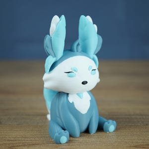 A studio photograph on a light wood grain surface and a grey-blue background shows the Winter Flutterfox miniature in light blue, dark blue, and white colors.