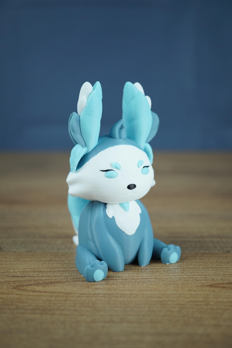 A studio photograph on a light wood grain surface and a grey-blue background shows the Winter Flutterfox miniature in light blue, dark blue, and white colors.