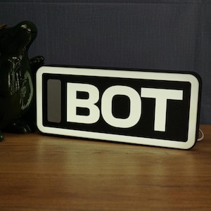 May include: A rectangular black and white sign displaying the word "BOT" in bold white letters. The sign has a white border and is on a wooden surface. A dark green decorative object is visible on the left.
