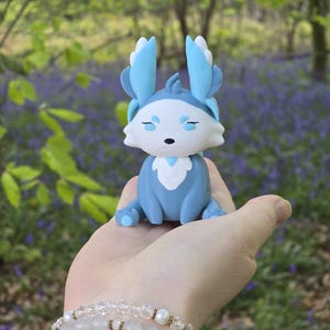 A hand-held shot featuring the Winter Flutterfox miniature, which has light blue, medium blue, and white colors, sitting on a palm against a blurred background of green forest leaves and purple bluebells.