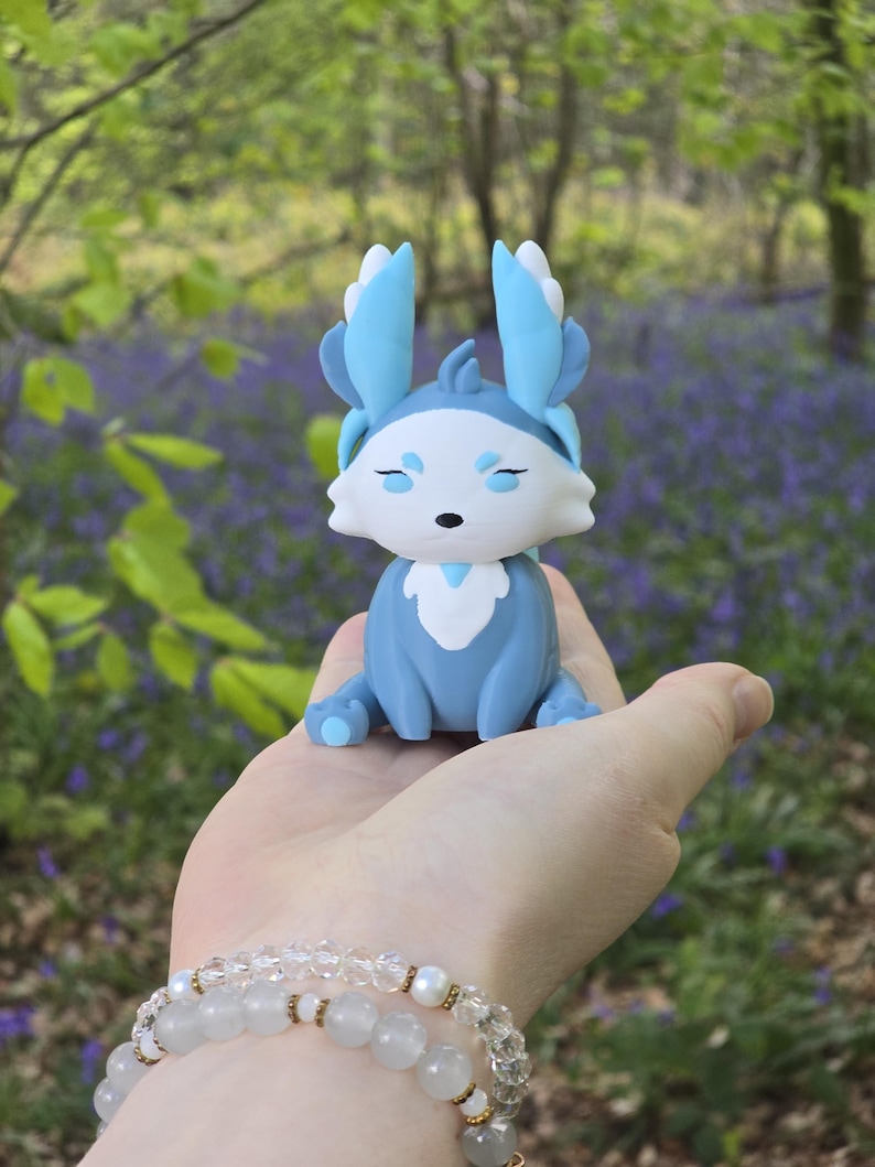 A hand-held shot featuring the Winter Flutterfox miniature, which has light blue, medium blue, and white colors, sitting on a palm against a blurred background of green forest leaves and purple bluebells.