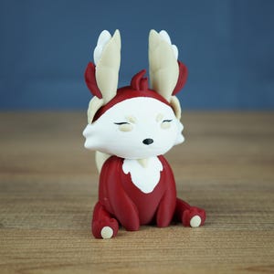 A studio product shot showing the Autumn Flutterfox miniature, which is dark red and white with cream and gold accents, sitting centered on a wooden surface with a soft grey-blue background.