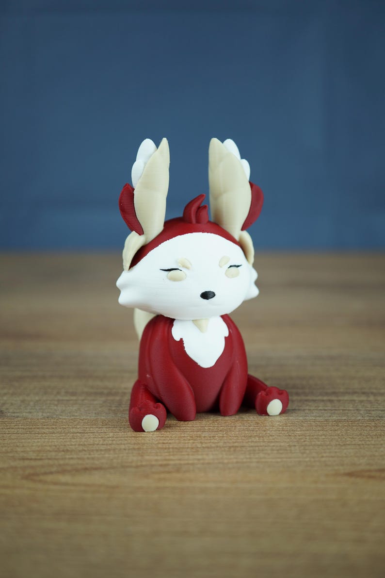 A studio product shot showing the Autumn Flutterfox miniature, which is dark red and white with cream and gold accents, sitting centered on a wooden surface with a soft grey-blue background.