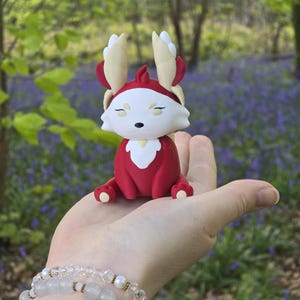 The Leafy Flutterfox miniature sits on a human hand. This figure features deep scarlet red and creamy white colors with gold accents, set against an outdoor bluebell and woodland background.