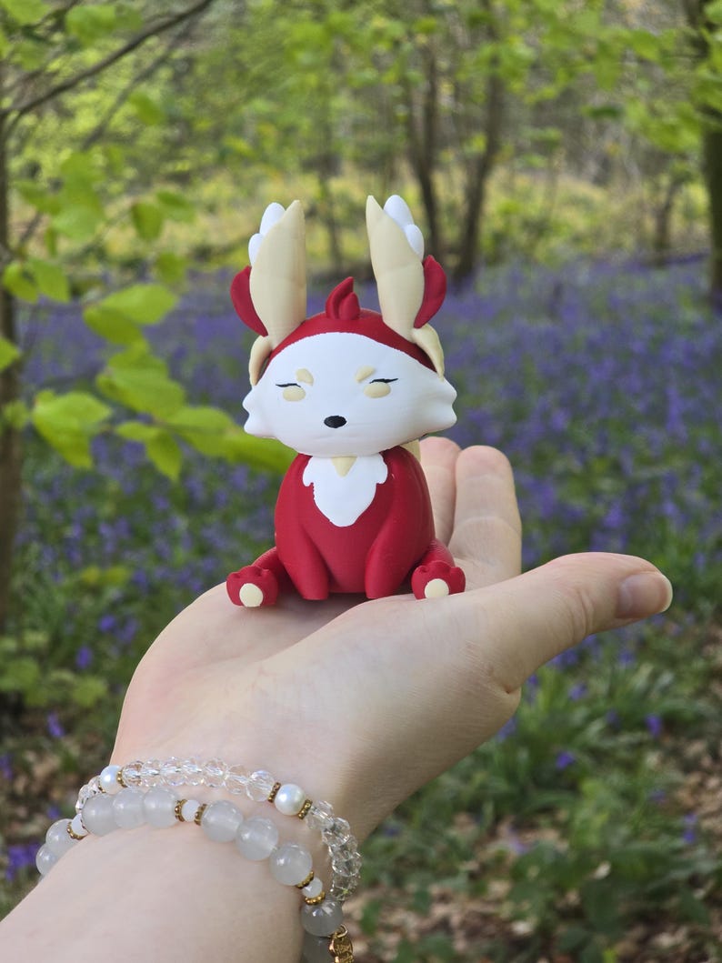 The Leafy Flutterfox miniature sits on a human hand. This figure features deep scarlet red and creamy white colors with gold accents, set against an outdoor bluebell and woodland background.