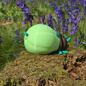 A high-angle, slightly-from-the-right three-quarter view of the 3D printed Palia Lantern Bug collectible. Positioned on a light wood surface, this perspective provides a clear look at the bug's brown striped back, fanned turquoise legs, and the detailed head with its black eyes and turquoise antennae. The massive, light-green glowing abdomen is prominently displayed, contrasting with the dark blue fabric background in the distance.