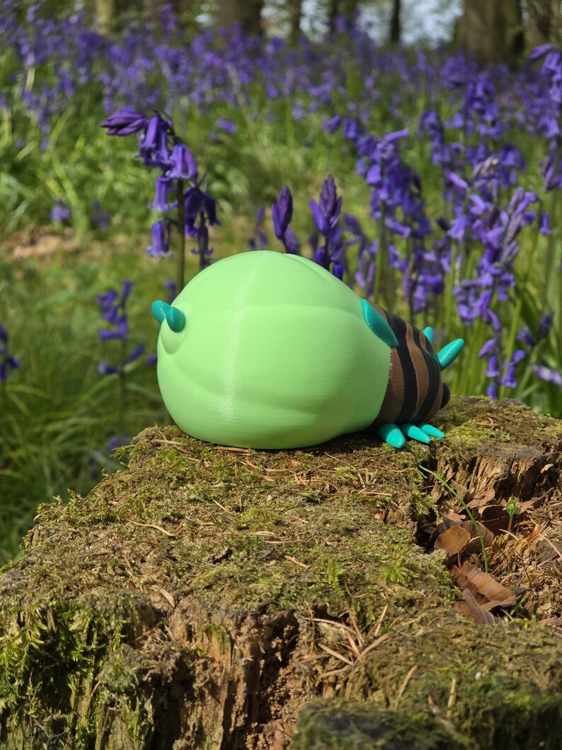 A high-angle, slightly-from-the-right three-quarter view of the 3D printed Palia Lantern Bug collectible. Positioned on a light wood surface, this perspective provides a clear look at the bug's brown striped back, fanned turquoise legs, and the detailed head with its black eyes and turquoise antennae. The massive, light-green glowing abdomen is prominently displayed, contrasting with the dark blue fabric background in the distance.