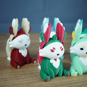 An angled close-up focusing on the Summer Flutterfox miniature, which is teal-green and white with bright pinkish-red leaf accents, sitting on a wooden surface. Other Flutterfox variants, like the red Autumn fox, are softly blurred in the background.