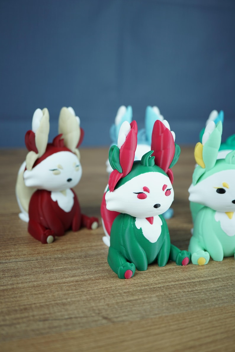 An angled close-up focusing on the Summer Flutterfox miniature, which is teal-green and white with bright pinkish-red leaf accents, sitting on a wooden surface. Other Flutterfox variants, like the red Autumn fox, are softly blurred in the background.