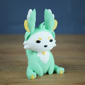 A product photo of the Spring Flutterfox miniature (mint green, white, and gold) sitting on a wooden table, against a diffused grey-blue studio backdrop.