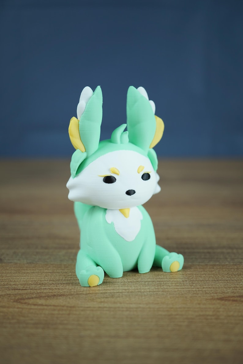 A product photo of the Spring Flutterfox miniature (mint green, white, and gold) sitting on a wooden table, against a diffused grey-blue studio backdrop.