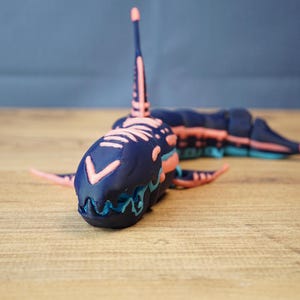 Op de afbeelding: Een gedetailleerde, 3D-geprinte speelgoedhaai in een levendig kleurenschema. De haai heeft een donkerblauwe body met roze en lichtblauwe accenten. Het ontwerp omvat vinnen, een rugvin en ingewikkelde patronen.