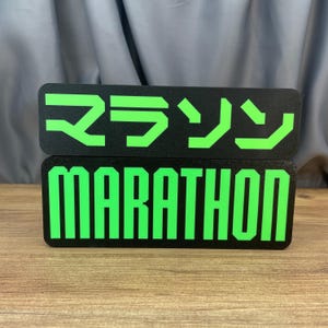 May include: Two black rectangular signs with green text. The top sign has Japanese characters. The bottom sign reads "MARATHON" in bold, block letters. The signs are likely for a marathon event.