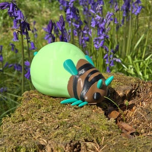 A high-quality 3D printed collectible figurine of the Palia Lantern Bug, resting on a light wood grain surface. The figure features a detailed brown head with black eyes and turquoise antennae, a black-and-brown striped segmented body, turquoise legs, and a large, smooth, light-green abdomen that glows in the dark. The pose is a dynamic, slightly turned three-quarter view, showcasing all its main features against a soft, blurred blue fabric background.