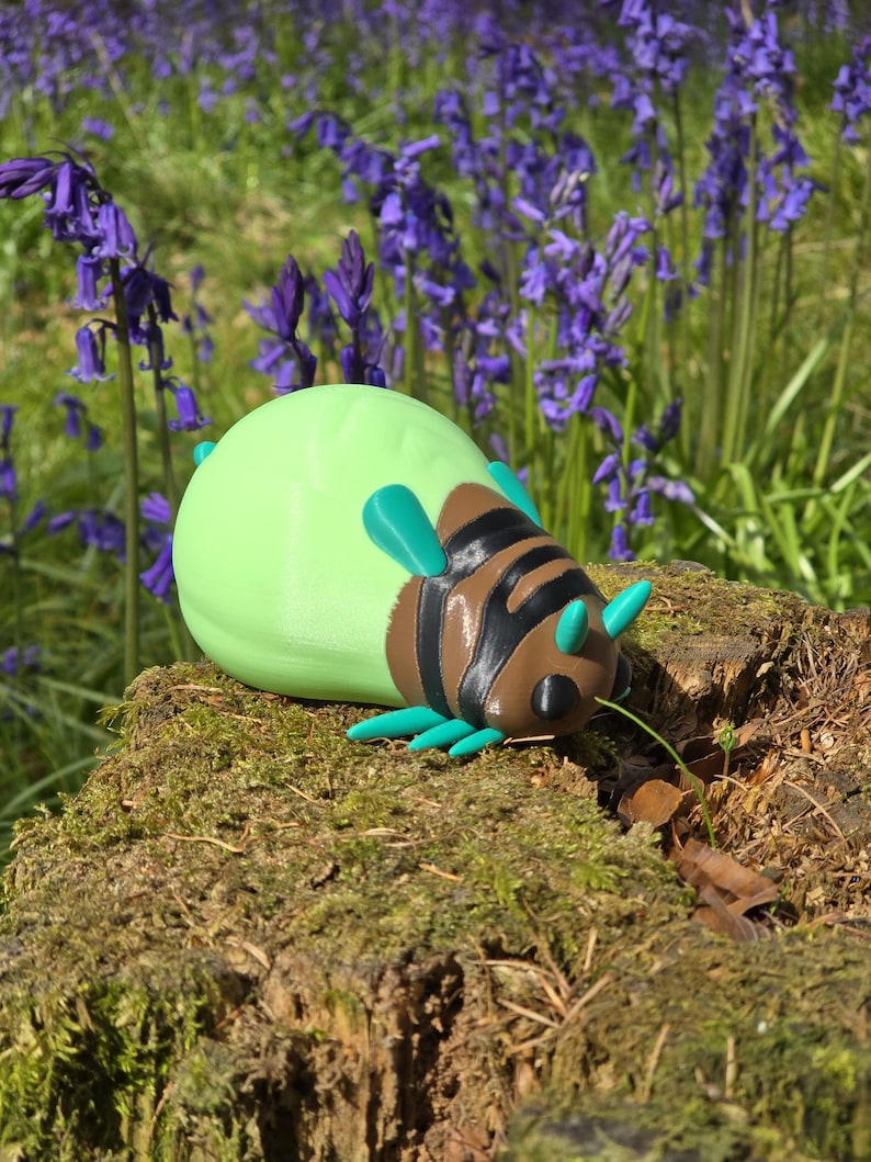 A high-quality 3D printed collectible figurine of the Palia Lantern Bug, resting on a light wood grain surface. The figure features a detailed brown head with black eyes and turquoise antennae, a black-and-brown striped segmented body, turquoise legs, and a large, smooth, light-green abdomen that glows in the dark. The pose is a dynamic, slightly turned three-quarter view, showcasing all its main features against a soft, blurred blue fabric background.