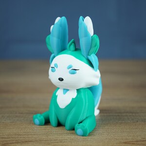 A product shot featuring the Fall Flutterfox miniature, which is deep teal and white with bright blue and green accents, sitting centrally on a wooden table.