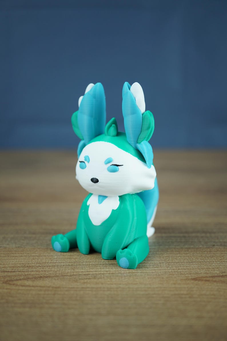A product shot featuring the Fall Flutterfox miniature, which is deep teal and white with bright blue and green accents, sitting centrally on a wooden table.
