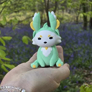 close-up hand photograph in a lush forest with bluebell flowers. It shows the Spring Flutterfox 3D-printed miniature (5cm or 9cm) sitting on a palm. The figure is light green and white with yellow leaf-like accents. A pearl and crystal bead bracelet is visible on the wrist.