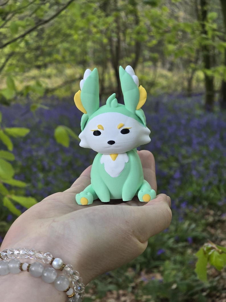 close-up hand photograph in a lush forest with bluebell flowers. It shows the Spring Flutterfox 3D-printed miniature (5cm or 9cm) sitting on a palm. The figure is light green and white with yellow leaf-like accents. A pearl and crystal bead bracelet is visible on the wrist.