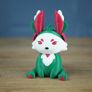 A studio photograph on a light wooden table against a soft blue-grey backdrop. The Summer Flutterfox miniature, with deep green and white body and bright pinkish-red leaf ears, sits facing forward.
