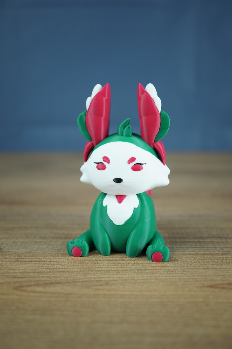 A studio photograph on a light wooden table against a soft blue-grey backdrop. The Summer Flutterfox miniature, with deep green and white body and bright pinkish-red leaf ears, sits facing forward.