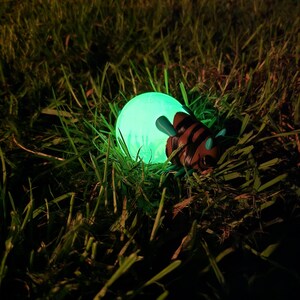 A captivating, real-world nighttime photograph of the 3D printed Palia Lantern Bug collectible glowing intensely within a patch of dark green grass. The figurine's light-green abdomen emits a powerful, enchanting green light, brightly illuminating the individual blades of grass and weeds surrounding it. The detailed brown head and turquoise antennae are partially visible against the glow. This shot, taken outdoors at night, perfectly captures the magical and atmospheric quality