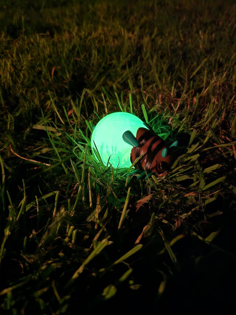 A captivating, real-world nighttime photograph of the 3D printed Palia Lantern Bug collectible glowing intensely within a patch of dark green grass. The figurine's light-green abdomen emits a powerful, enchanting green light, brightly illuminating the individual blades of grass and weeds surrounding it. The detailed brown head and turquoise antennae are partially visible against the glow. This shot, taken outdoors at night, perfectly captures the magical and atmospheric quality