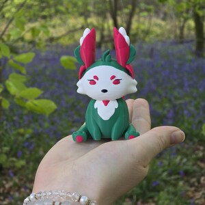A human palm, complete with a crystal and pearl bracelet, holds the Summer Flutterfox miniature. This figure is primarily deep teal-green and white with bright pinkish-red leaf accents, photographed outdoors with bluebells in the background.