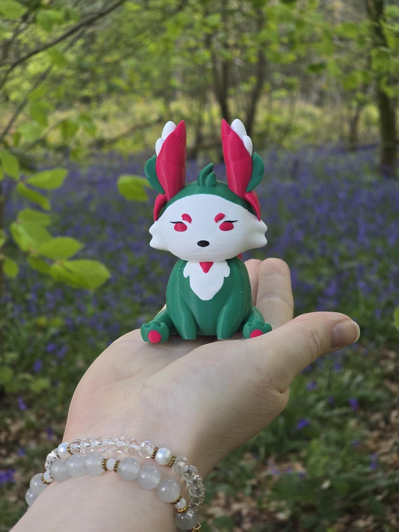 A human palm, complete with a crystal and pearl bracelet, holds the Summer Flutterfox miniature. This figure is primarily deep teal-green and white with bright pinkish-red leaf accents, photographed outdoors with bluebells in the background.