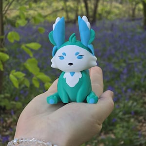 A close-up view of the Fall Flutterfox 3D-printed miniature, sitting on a hand, against a backdrop of natural forest and bluebells. The figure has a rich turquoise and dark red color scheme.